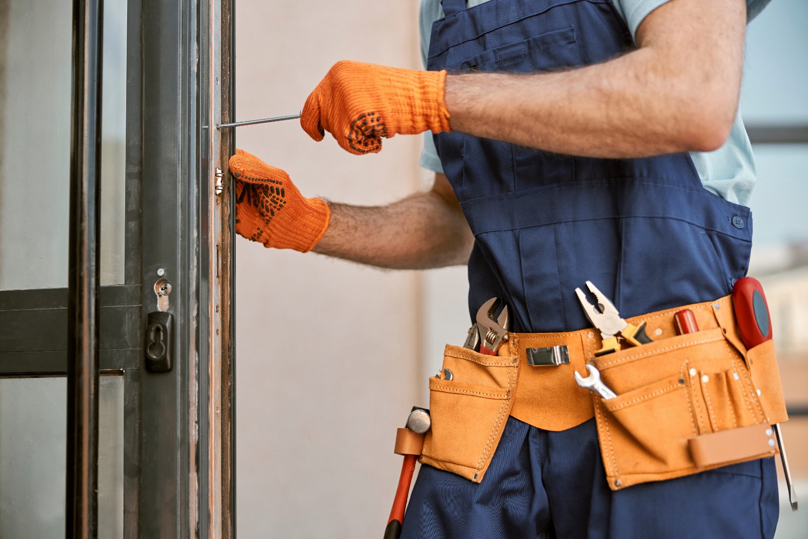 Tradesman in orange gloves adjusting door lock with screwdriver and tool belt